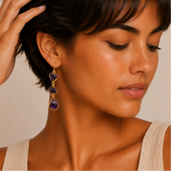 Close-up of a woman wearing gold and purple dangling earrings against a neutral background