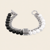 A silver-toned chain bracelet featuring one side with matte black beads and the other with white marbled beads, finished with a textured central cap and toggle clasp, shown on a light beige background.