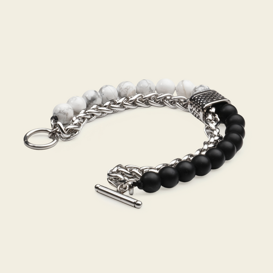 A men's multi-strand bracelet featuring silver-tone chain links, a textured silver spacer, white marbled beads, and matte black round beads, finished with a toggle clasp on a light beige background.