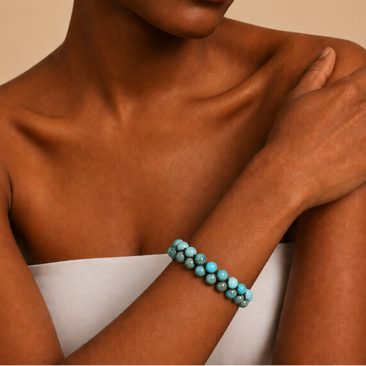 A close-up of a model wearing a double-strand turquoise beaded bracelet on her wrist, with her arms crossed over her chest against a warm beige background.