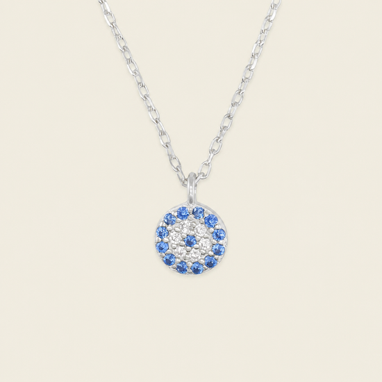 A silver chain necklace with a circular pendant featuring a blue and white eye design, set against a solid light beige background.