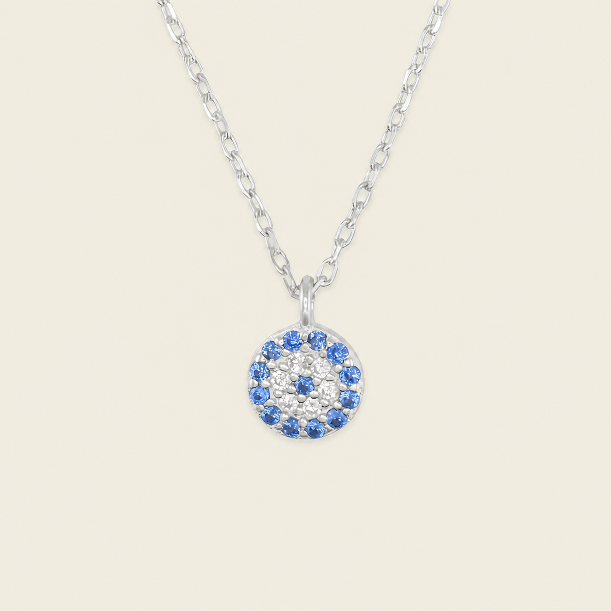 A silver chain necklace with a circular pendant featuring a blue and white eye design, set against a solid light beige background.