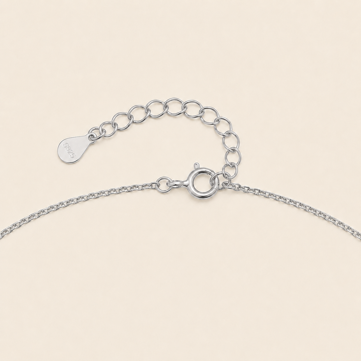 Close-up of a silver chain necklace showing a lobster clasp, extension chain, and a small engraved teardrop tag on a light beige background.