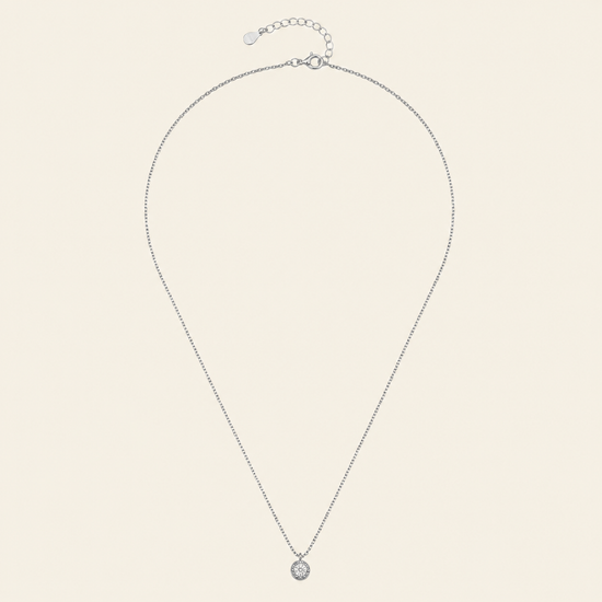 A delicate silver chain necklace with a small round bezel-set clear gemstone pendant, displayed on a light cream background.