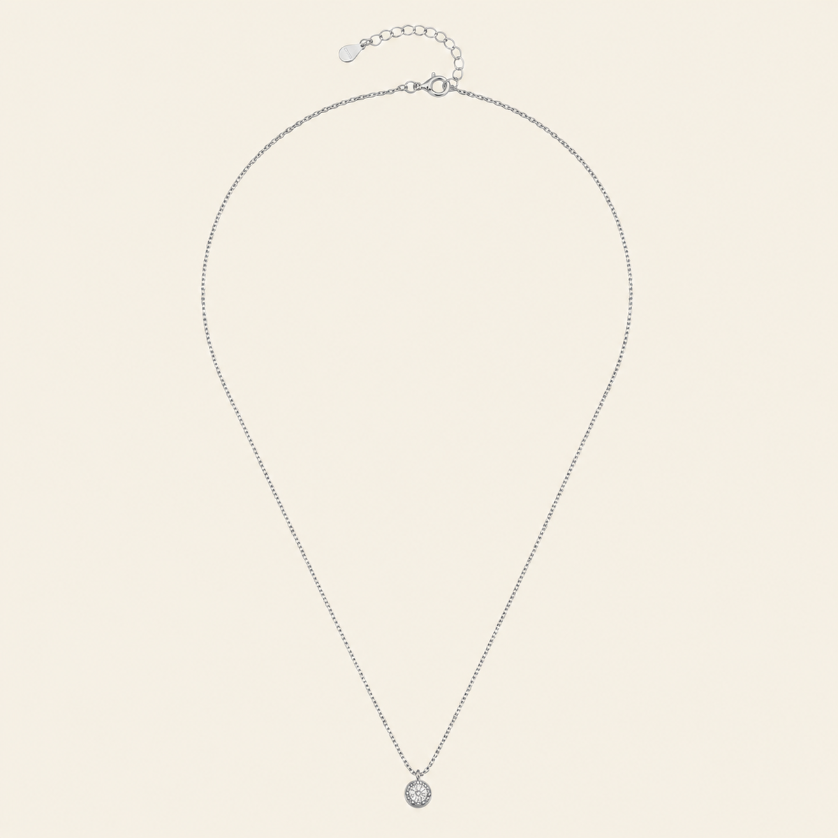A delicate silver chain necklace with a small round bezel-set clear gemstone pendant, displayed on a light cream background.