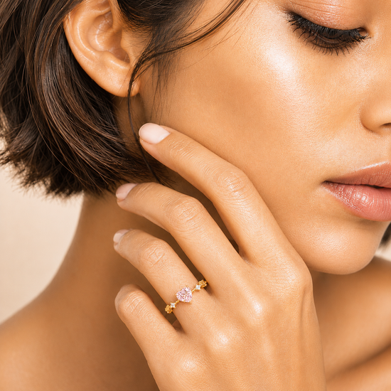 A close-up lifestyle shot of a model wearing a delicate gold ring with a small pink heart-shaped gemstone on her finger.