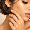 A close-up lifestyle shot of a model wearing a delicate gold ring with a small pink heart-shaped gemstone on her finger.