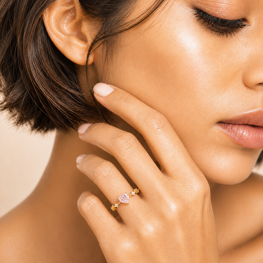 A close-up lifestyle shot of a model wearing a delicate gold ring with a small pink heart-shaped gemstone on her finger.