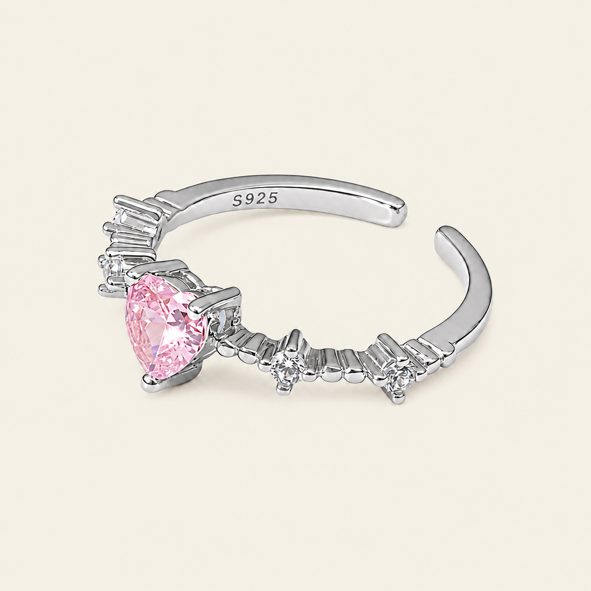 A silver-toned ring with a faceted pink heart-shaped center stone and small clear accent stones, shown on a plain light beige background. The inner band is marked S925.