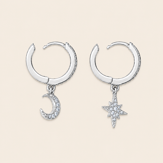 A pair of silver crescent moon and star hoop earrings with clear crystal accents, displayed on a light beige background.