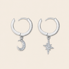 A pair of silver crescent moon and star hoop earrings with clear crystal accents, displayed on a light beige background.