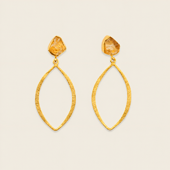 A pair of gold dangle earrings with textured open teardrop-shaped hoops and rough, faceted amber-colored crystal studs, shown against a solid light beige background.