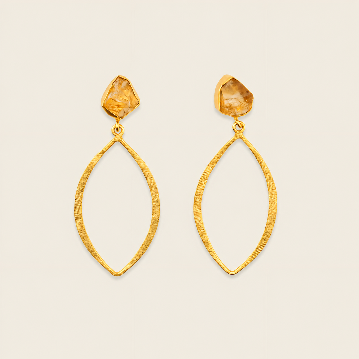 A pair of gold dangle earrings with textured open teardrop-shaped hoops and rough, faceted amber-colored crystal studs, shown against a solid light beige background.