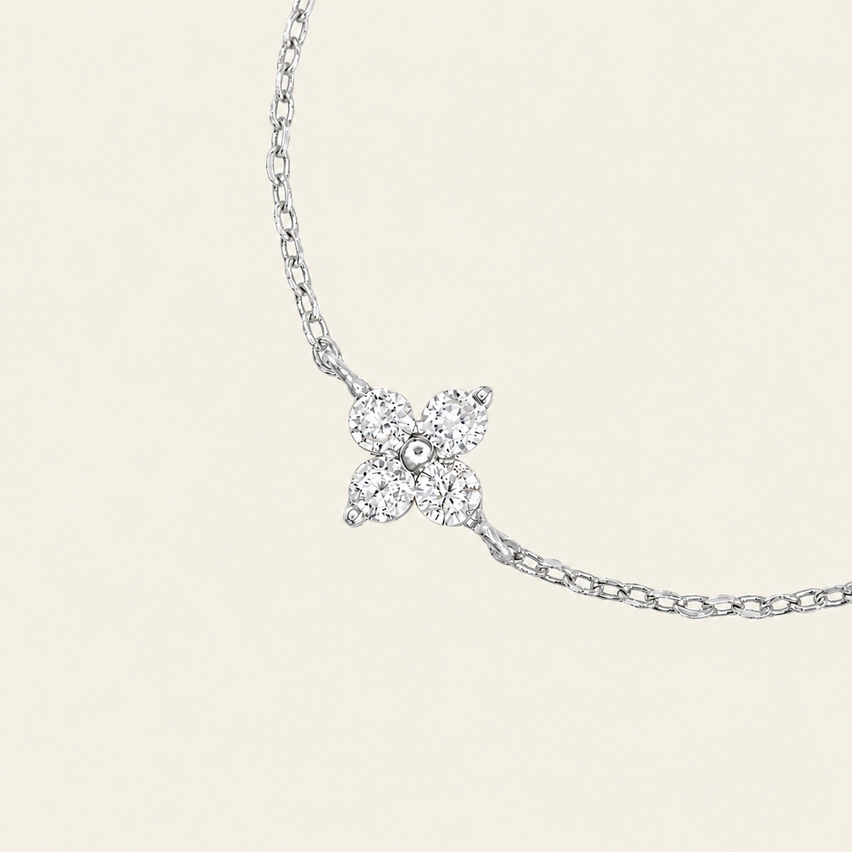 A delicate silver-toned chain bracelet featuring a small four-petal flower pendant set with clear stones, shown against a solid light beige background.