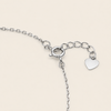 Silver chain bracelet detail showing a lobster clasp, extension chain, and a small polished heart charm on a light beige background.