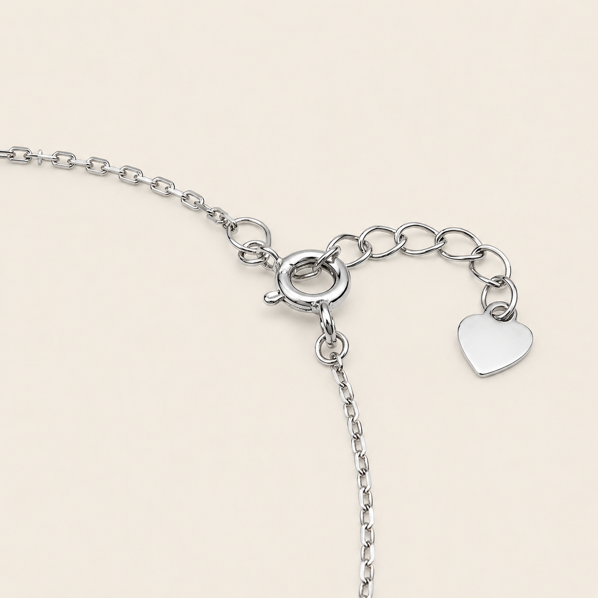 Silver chain bracelet detail showing a lobster clasp, extension chain, and a small polished heart charm on a light beige background.