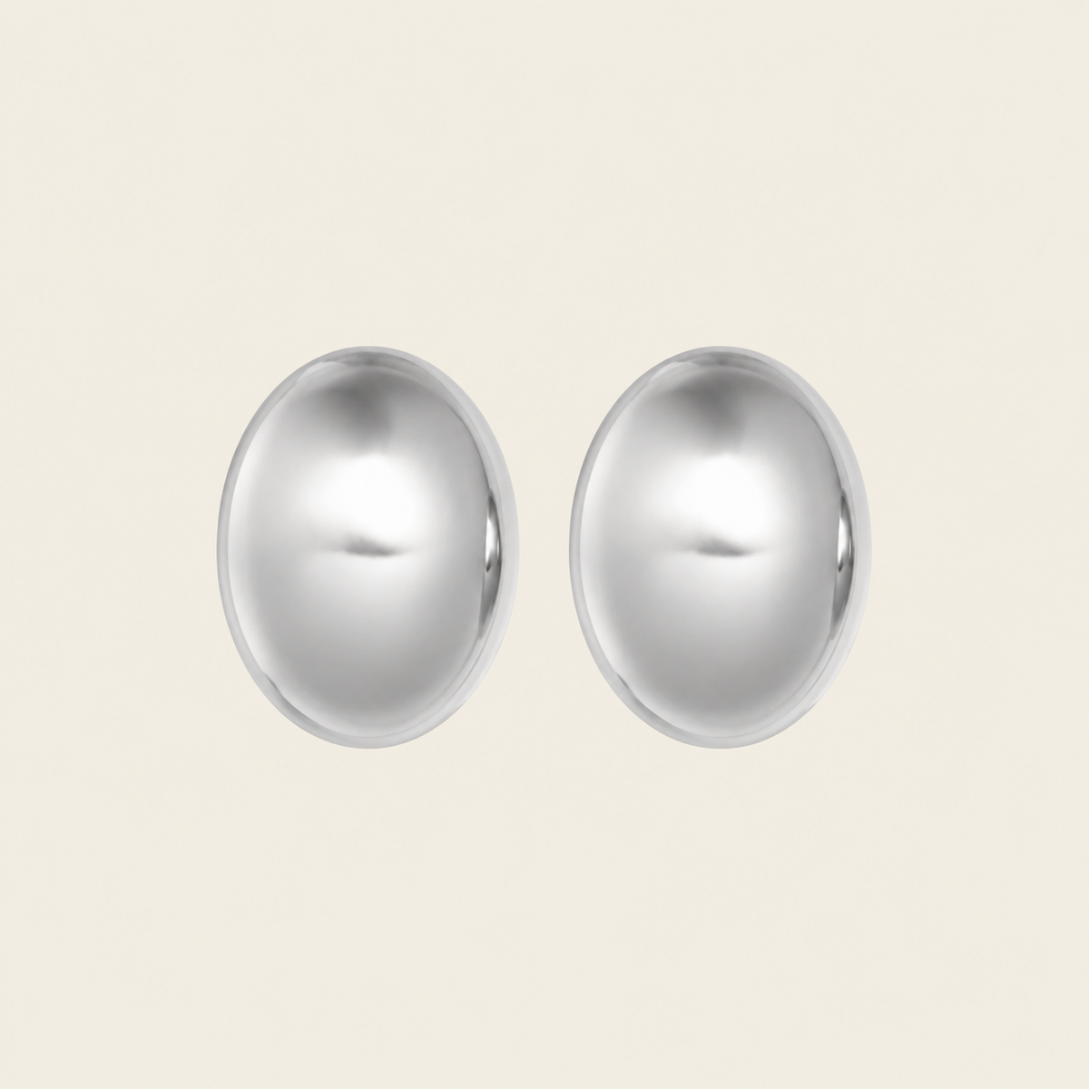 A pair of oval stud earrings with a polished, reflective silver finish, displayed against a solid, warm off-white background.