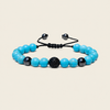 A beaded stretch bracelet featuring bright turquoise-blue beads, black marbled beads, and a central textured black sphere, strung on a black adjustable cord with black metal slider ends, set against a solid light beige background.