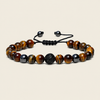 A beaded bracelet made of polished tiger's eye beads in brown and black, with a textured black central bead and a black adjustable cord closure, shown on a light beige background.