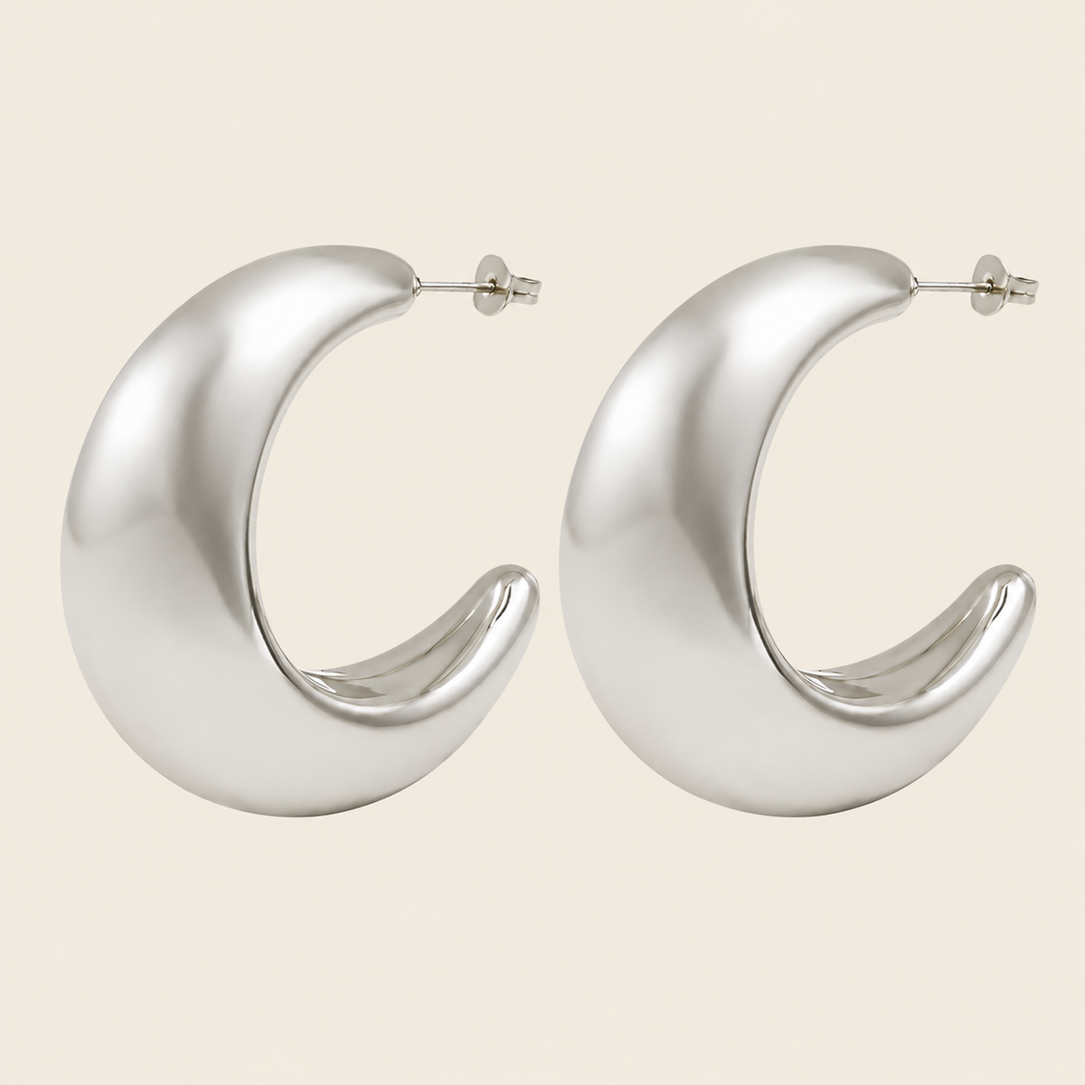 A pair of large, polished silver crescent-shaped hoop earrings on a solid light beige background.
