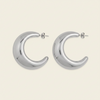 A pair of polished silver crescent-shaped hoop earrings with post backings, displayed on a light cream background.