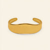 A polished gold cuff bracelet with a smooth, wavy profile, displayed on a clean, solid cream-colored background.