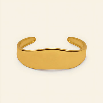 A polished gold cuff bracelet with a smooth, wavy profile, displayed on a clean, solid cream-colored background.
