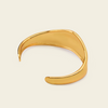 A polished gold-toned open cuff bangle bracelet with a smooth, organic wavy curve, displayed on a light cream background.