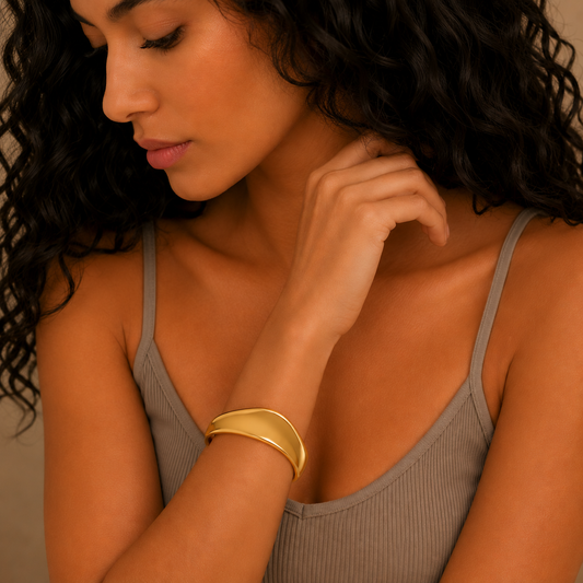 A close-up lifestyle shot of a woman wearing a wide, polished gold cuff bracelet on her wrist, paired with a ribbed taupe tank top.