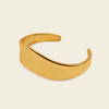 A polished gold-toned metal cuff bracelet with a smooth, curved, open design, displayed on a solid light cream background.