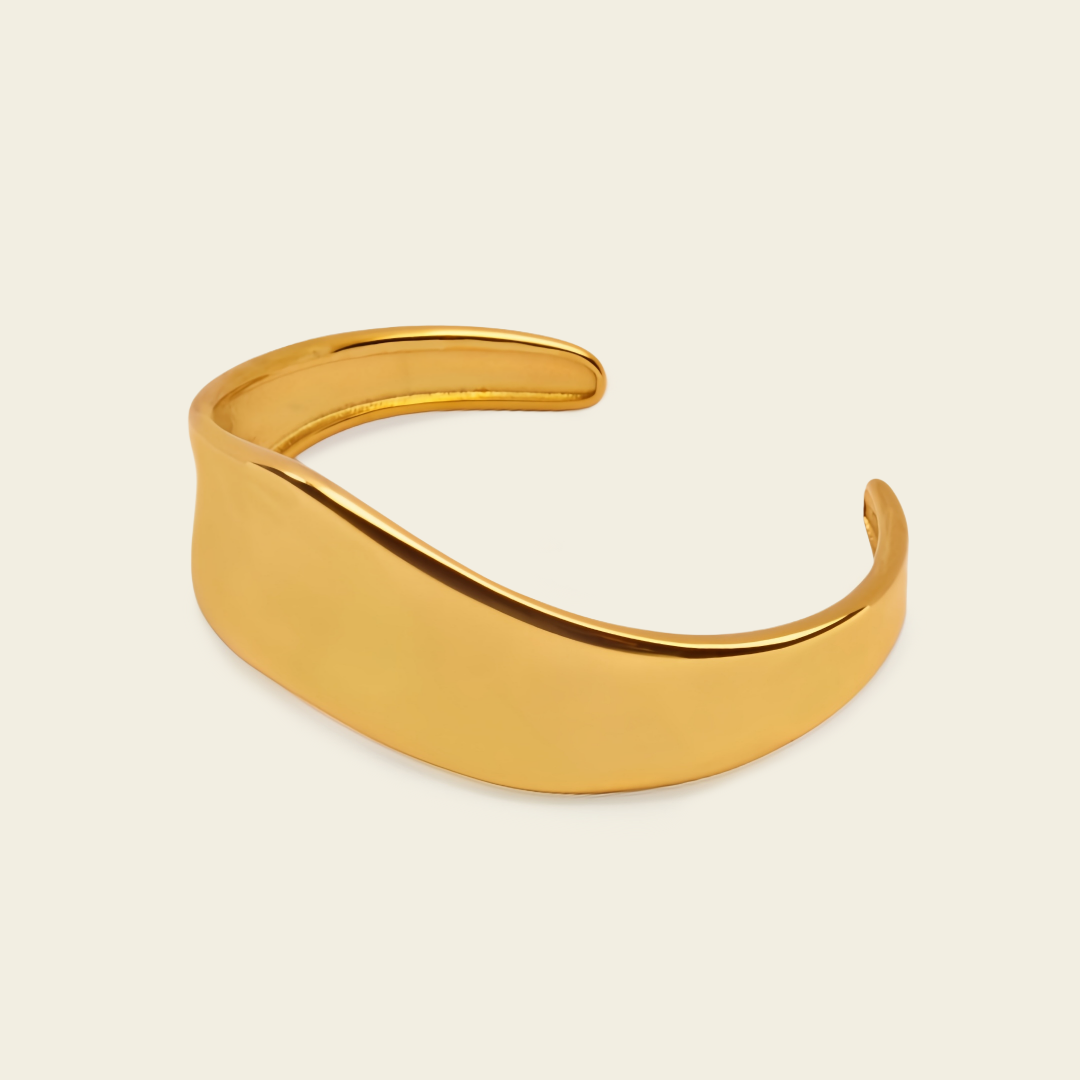 A polished gold-toned metal cuff bracelet with a smooth, curved, open design, displayed on a solid light cream background.