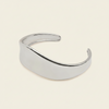 A polished silver-toned metal cuff bracelet with a smooth, curved, open design, displayed on a solid light cream background.