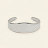 A polished silver cuff bracelet with a smooth, wavy profile, displayed on a clean, solid cream-colored background.