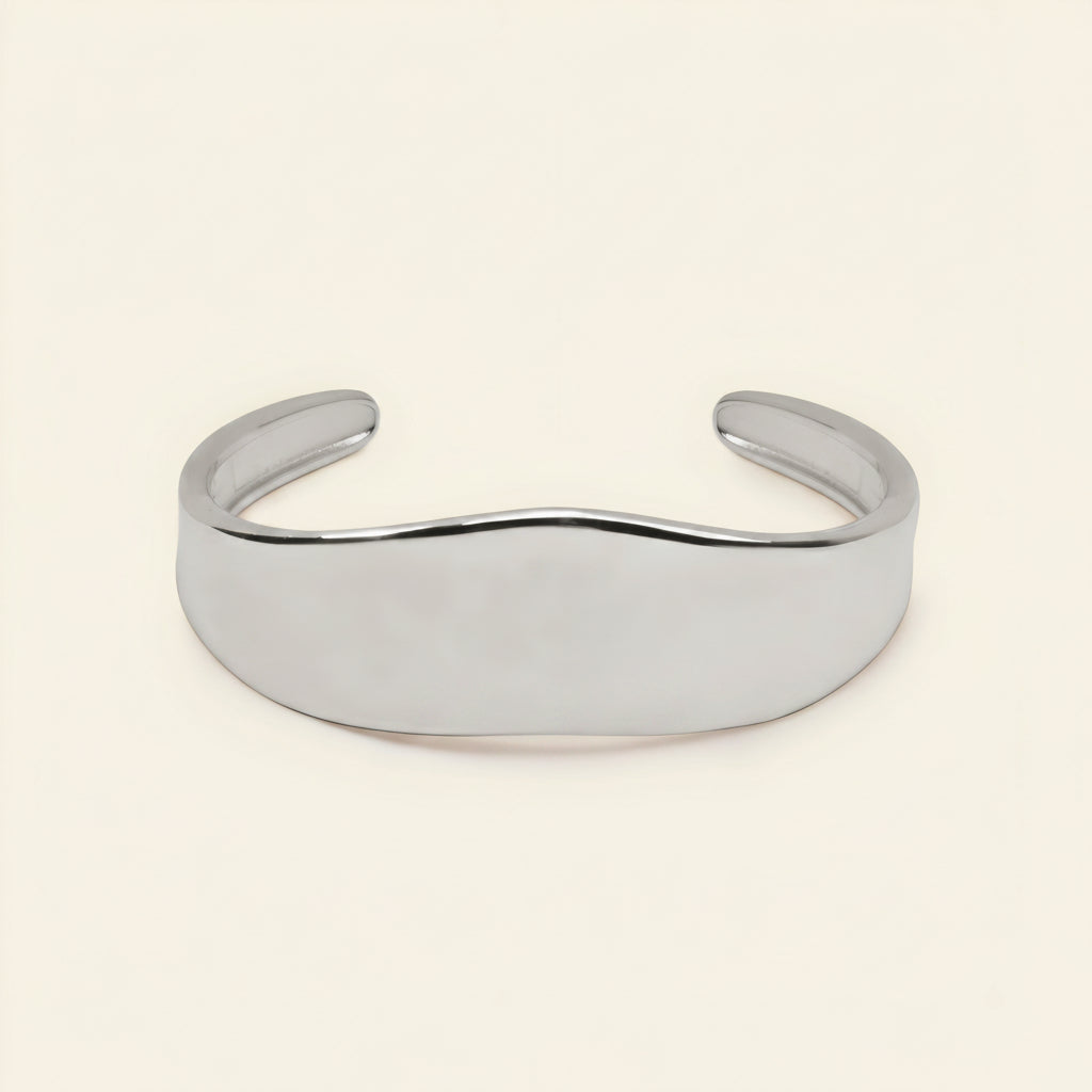 A polished silver cuff bracelet with a smooth, wavy profile, displayed on a clean, solid cream-colored background.