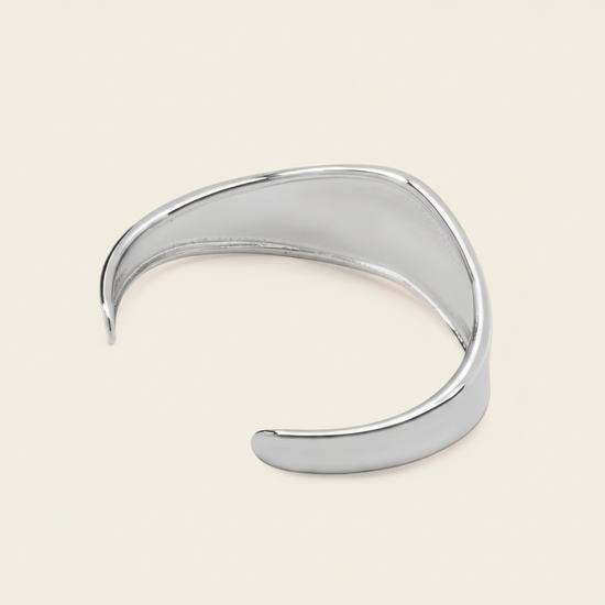 A polished silver-toned open cuff bangle bracelet with a smooth, organic wavy curve, displayed on a light cream background.