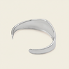 A polished silver-toned open cuff bangle bracelet with a smooth, organic wavy curve, displayed on a light cream background.