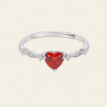 A silver-toned adjustable ring with a heart-shaped red gemstone in the center and small clear accent stones along the band, shown on a light cream background.