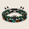 A set of two beaded stretch bracelets on a light beige background. The bracelets feature green marbled beads, turquoise beads, brown tiger's eye beads, black beads, and textured black metal spacer rings, with one bracelet featuring a central black crown charm.