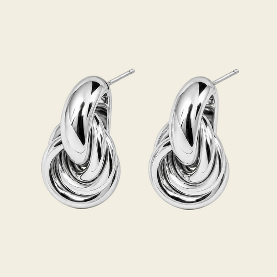 A pair of polished silver-toned earrings with a sculptural, intertwined knot-like design and post backs, displayed on a solid light beige background.