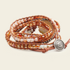A multi-strand wrap bracelet featuring brown leather cords, peach and cream beads, faceted amber glass beads, and a round silver-toned metal charm, displayed on a light beige background.