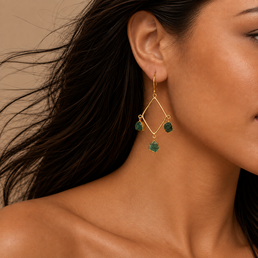 A close-up of a woman wearing a gold geometric dangle earring with three green gemstone drops, set against a warm beige background.