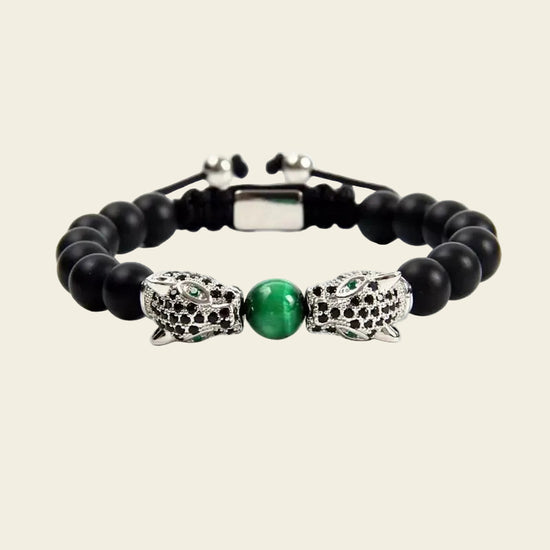 A beaded bracelet featuring matte black round beads, a central polished green oval stone, and two silver-toned panther head charms embellished with small black stones, shown on a light beige background.