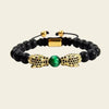 A beaded stretch bracelet featuring matte black round beads, two gold-toned crocodile or dragon head charms studded with small black stones, and a central green oval bead, shown on a light beige background.