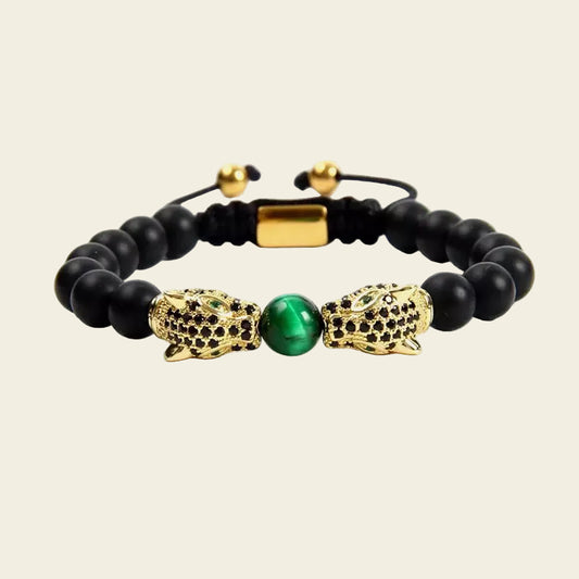A beaded stretch bracelet featuring matte black round beads, two gold-toned crocodile or dragon head charms studded with small black stones, and a central green oval bead, shown on a light beige background.