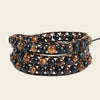 A multi-strand wrap bracelet featuring alternating glossy black and brown tiger's eye beads on a black cord, set against a solid light beige background.