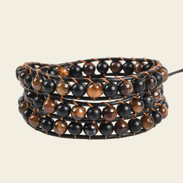 A multi-strand wrap bracelet featuring alternating glossy black and brown tiger's eye beads on a black cord, set against a solid light beige background.