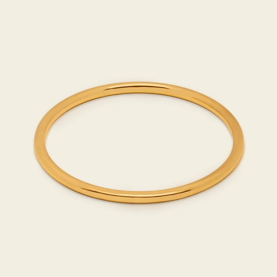 A thin, polished gold bangle bracelet shown on a clean, solid, light cream background.
