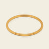 A thin, polished gold bangle bracelet shown on a clean, solid, light cream background.