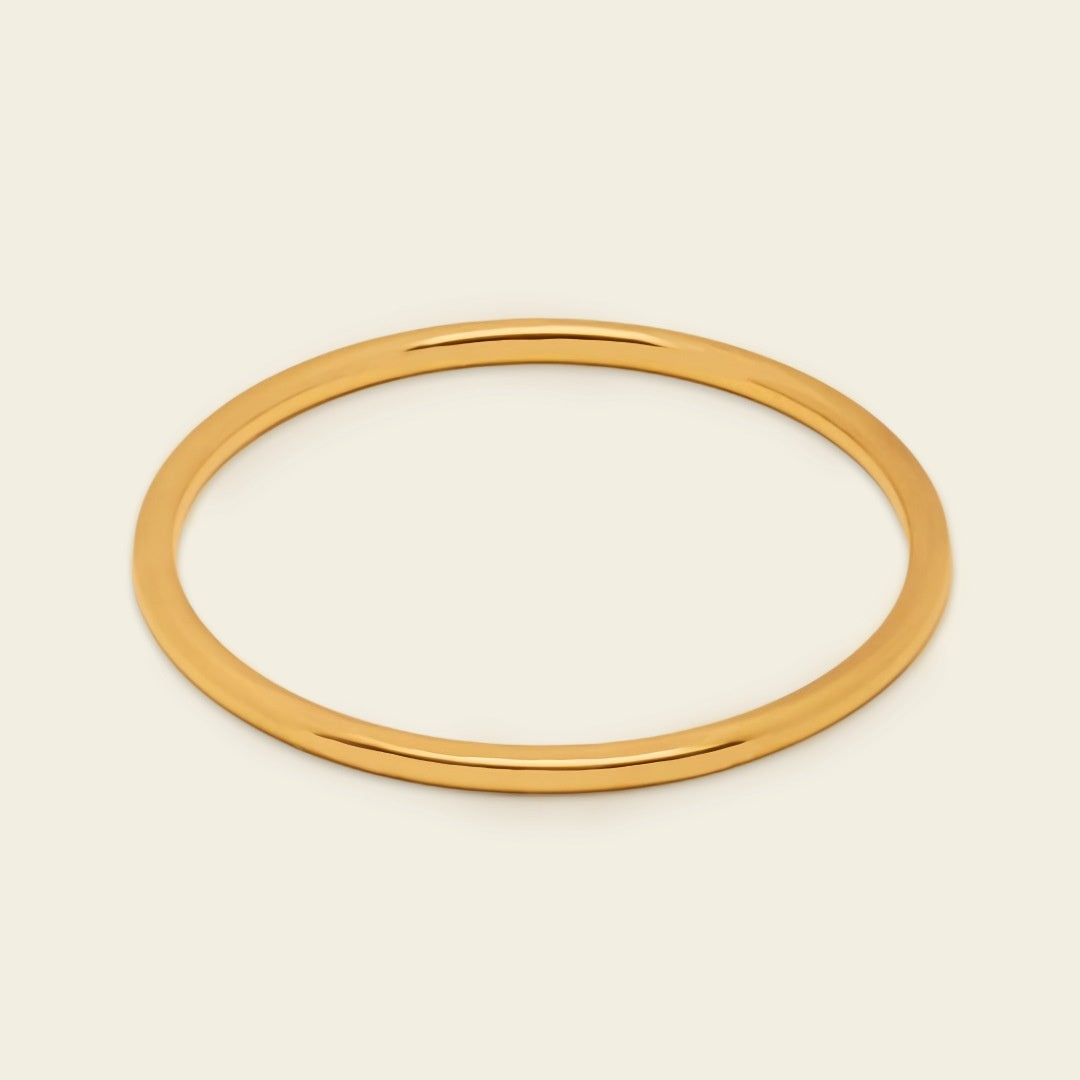A thin, polished gold bangle bracelet shown on a clean, solid, light cream background.