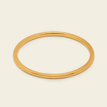 A thin, polished gold bangle bracelet shown on a clean, solid, light cream background.